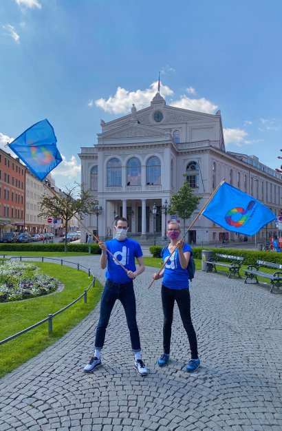 Zwei Mitglieder*innen von diversity München schwenken diversity-Fahnen am Gärtnerplatz in München anlässlich des IDAHOBIT* 2020.